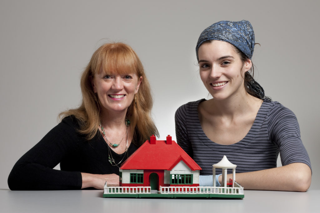 Two women sit at a table behind a toy house made of plastic construction bricks. It is a high-roofed single-storey house made up of red, green and white 'BAYKO' parts with a lawn, white fence, path and a small gazebo to the house's right. 